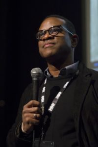 Thomas Allen Harris holds a microphone standing in front of a screen