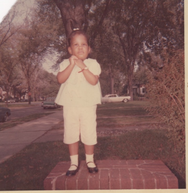 Small girl dressed in all white stands on brick walk way. Sidewalks in the background. Color photo