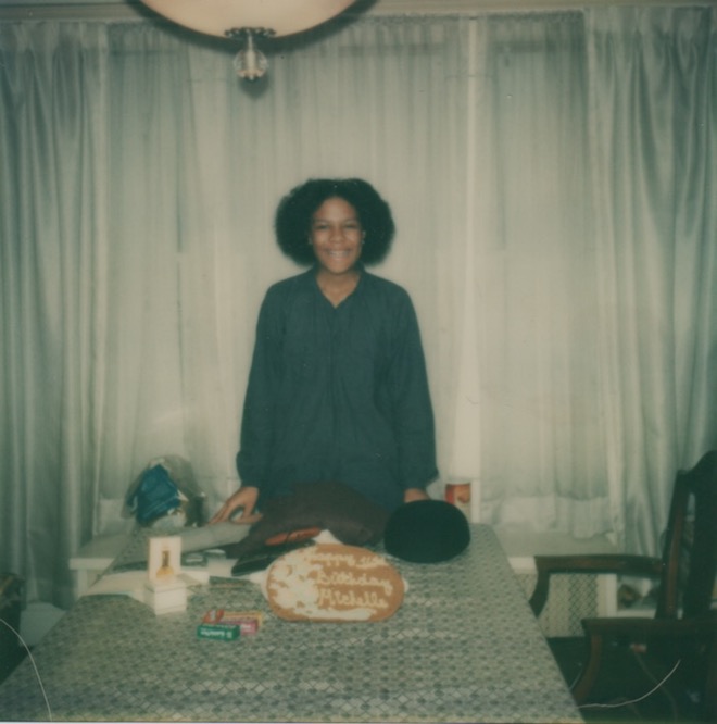 Young woman stands at the end of a table with a cake in front of her. She smiles at the camera. Color photo