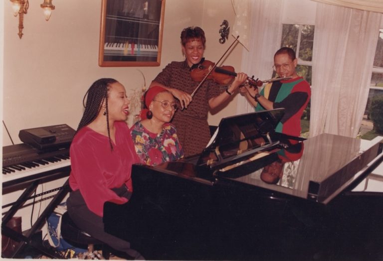 Two women sit at a piano. Two other individuals play violin to the site. Color Photo.