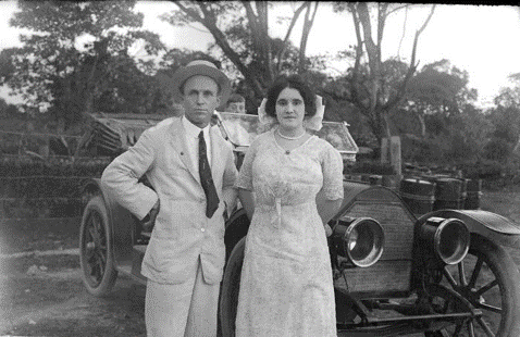 Robert and Aura pose in front of a car with straight faces while wearing a suit and day gown