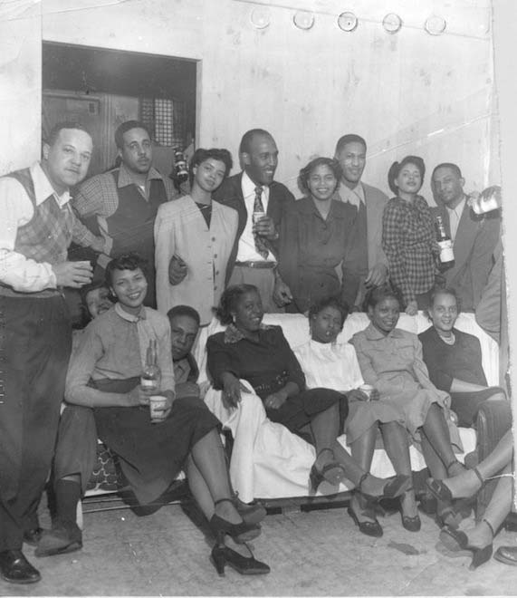 Group standing and sitting posed in the back of the Hastings St shop with Von Battles wife in plaid, and two of her five sisters, standing Von Battle is just out of camera